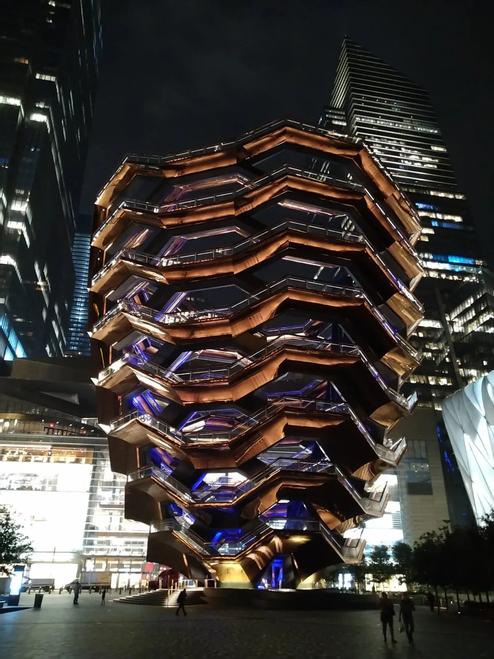 The image showcases a striking honeycomb-like structure illuminated at night with modern skyscrapers in the background highlighting a blend of innovative architecture and urban nightlife