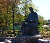 The image shows a statue of a seated person engrossed in reading a book set in a sunlit park with trees and a pedestrian in the background