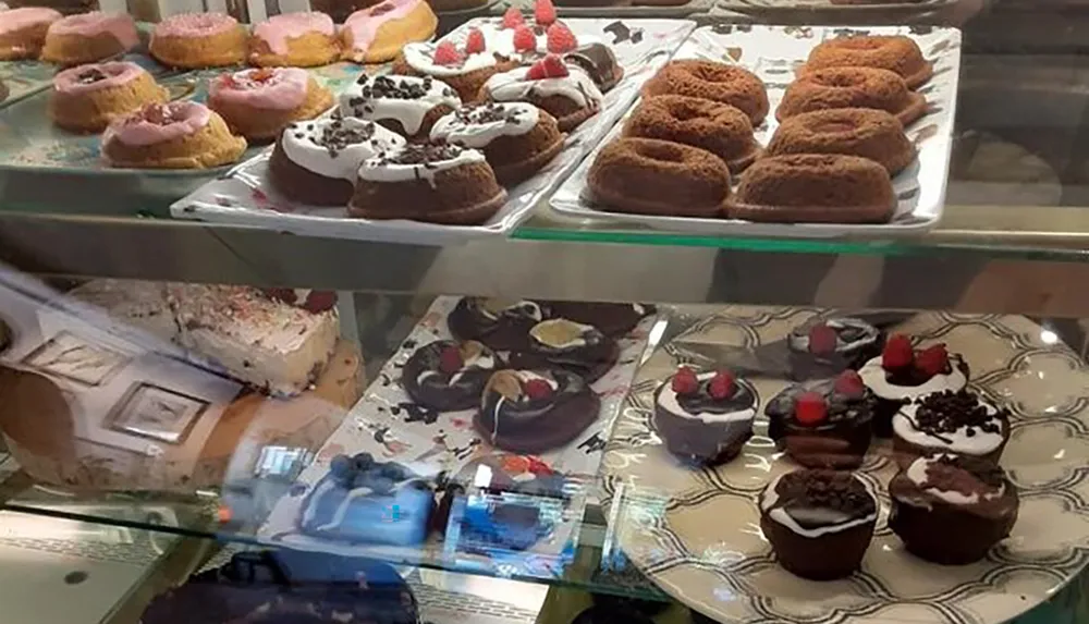 The image shows an assortment of frosted donuts bundt cakes and chocolate-covered desserts with strawberries showcased in a glass display case at a bakery or caf