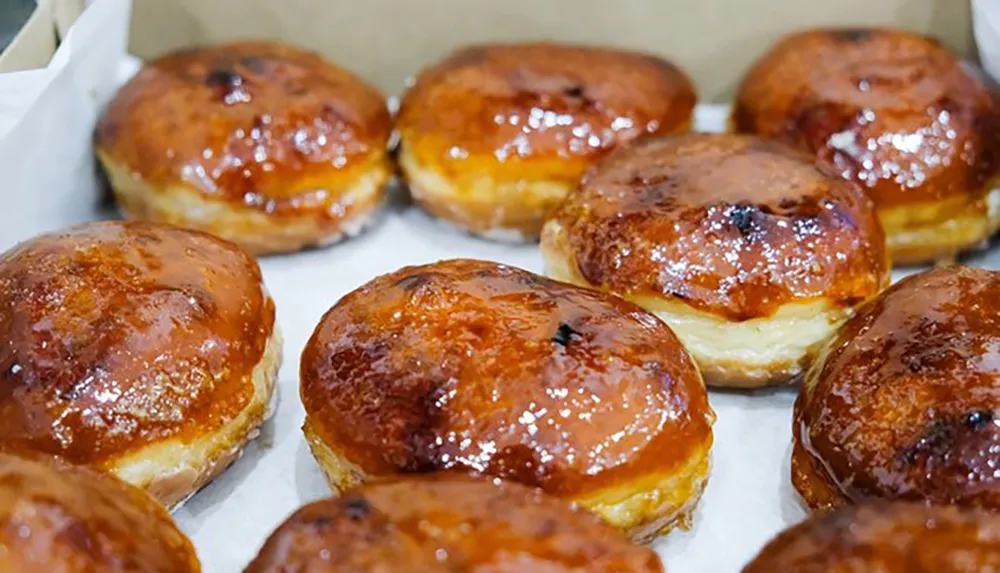 The image shows a box of glossy glazed jelly doughnuts with a golden brown crust suggesting they are freshly baked and coated with a sugary glaze