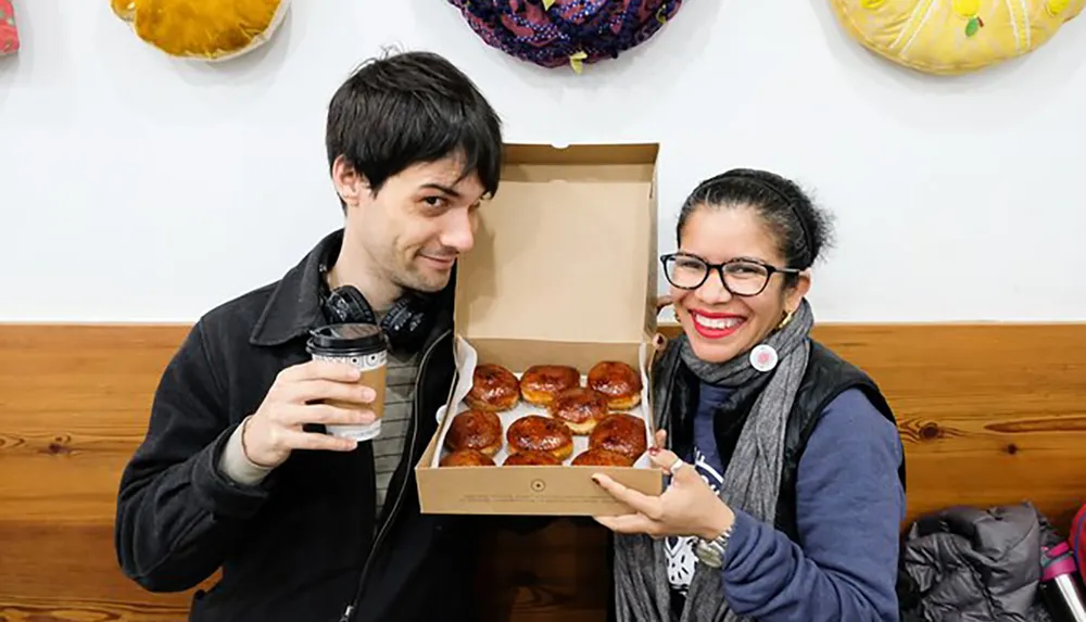 A man and a woman are smiling at the camera with the woman holding up a box of glazed pastries as the man holds a coffee cup both appearing cheerful and possibly celebrating a happy occasion or enjoying a casual treat