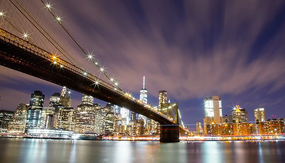 The image captures a long exposure night view of a lit suspension bridge with starburst effects on the lights stretching over a tranquil body of water towards a vibrant city skyline