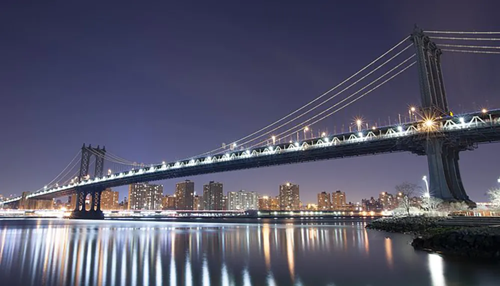 The image captures a beautifully illuminated suspension bridge at night reflecting over calm waters with a backdrop of city buildings