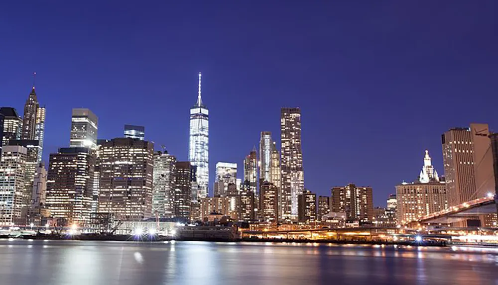 The image shows a stunning night view of the illuminated skyline of Lower Manhattan with the One World Trade Center prominently visible alongside calm waters and clear skies