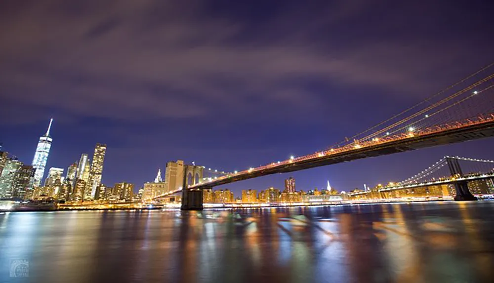 The photo captures the illuminated skyline of Manhattan at night with the Brooklyn Bridge spanning the foreground above a reflective body of water