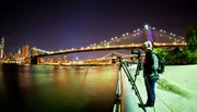 A person stands beside a camera on a tripod capturing a long-exposure photograph of a brightly lit suspension bridge at night, with a city skyline in the background.