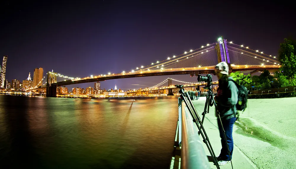 A person stands beside a camera on a tripod capturing a long-exposure photograph of a brightly lit suspension bridge at night with a city skyline in the background