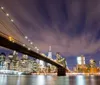 The image shows a long exposure night shot of the Brooklyn Bridge with radiant stars of light and streaky clouds above set against the backdrop of the illuminated New York City skyline