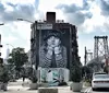 This image features a large building mural of a childs face with an urban street scene and the Williamsburg Bridge in the background