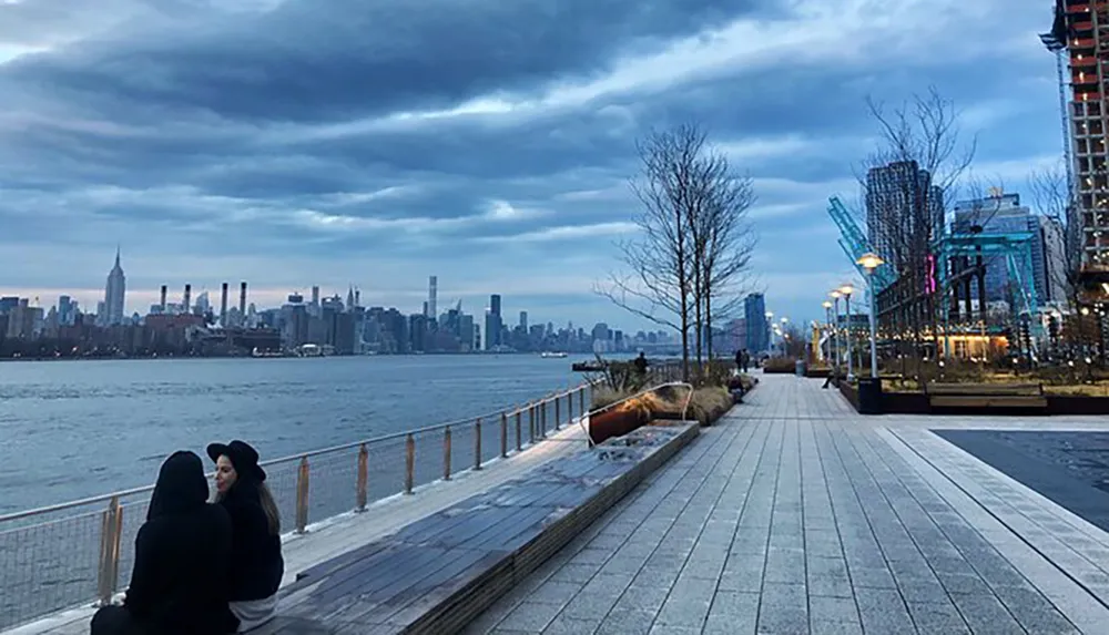 Two people are sitting on a waterfront boardwalk against the backdrop of a city skyline at dusk