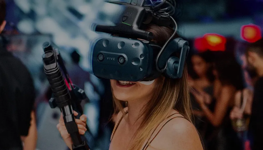 A person is smiling while wearing a virtual reality headset and holding a VR gun controller likely engaged in an immersive gaming experience