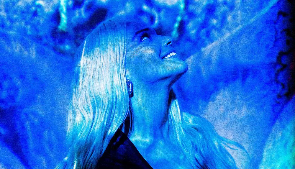 A woman with blonde hair is looking up with a smile in a room bathed in vibrant blue light