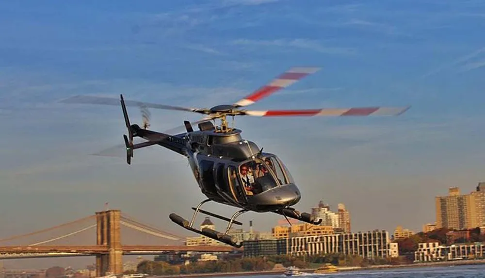 A helicopter is flying over a city skyline near a large bridge during what appears to be clear weather conditions