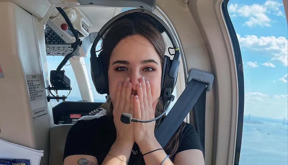 A person is sitting in the cockpit of a small aircraft wearing a headset and covering their mouth with their hands in apparent astonishment or excitement