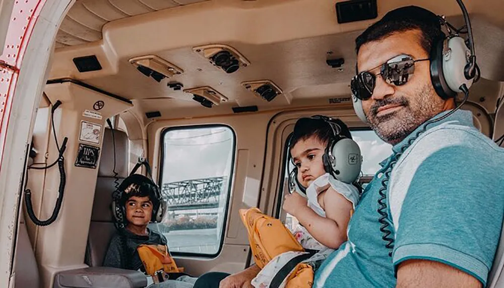 A person with two children all wearing headphones are seated inside a helicopter giving the impression of a family adventure