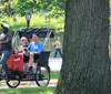 Two people are smiling while sitting in a red pedicab in an urban park setting with historic buildings in the background