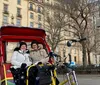 Two people are smiling while sitting in a red pedicab in an urban park setting with historic buildings in the background