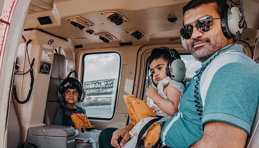 A man with sunglasses and two young children wearing headsets are seated inside a helicopter suggesting they are ready for a flight experience
