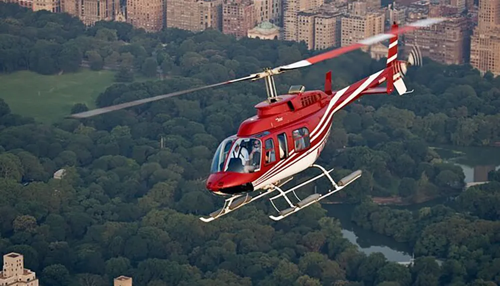 A red and white helicopter is flying over a large park surrounded by city buildings