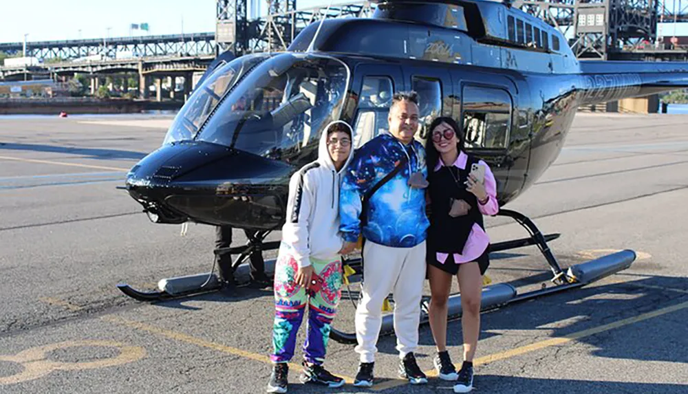 Three individuals are posing for a photo in front of a dark-colored helicopter on a sunny day with the backdrop featuring an industrial bridge structure