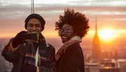A smiling man and woman are taking a selfie with a smartphone against a backdrop of a city skyline at sunset.