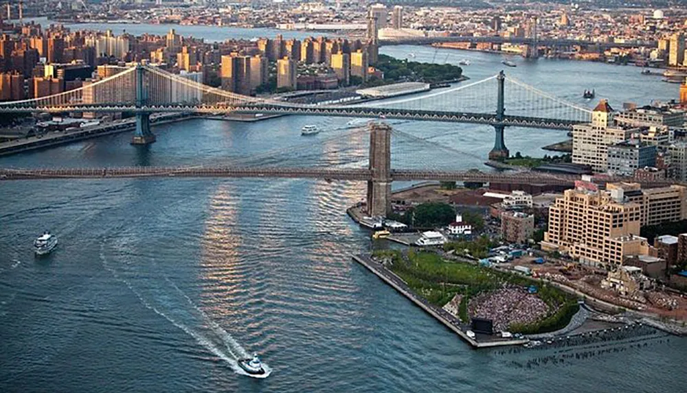 The image shows an aerial view of the East River in New York City with the Brooklyn and Manhattan Bridges connecting Brooklyn to Manhattan along with boats on the water and a crowded park area near the rivers edge