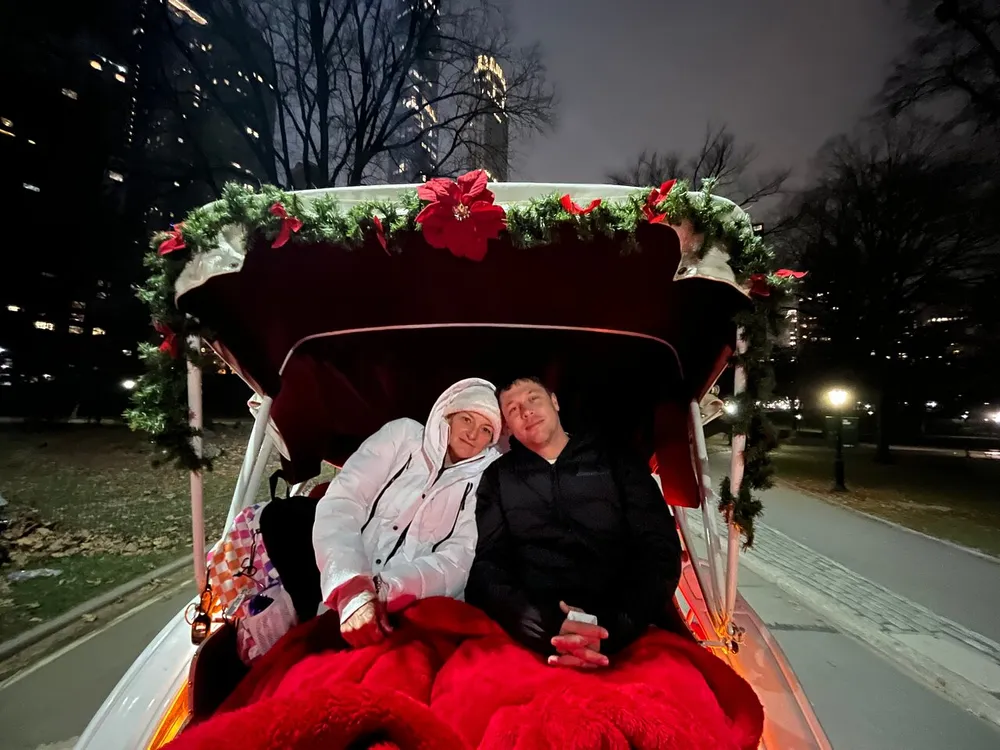 A couple is smiling while cozied up under a red blanket in a festively decorated horse-drawn carriage at night