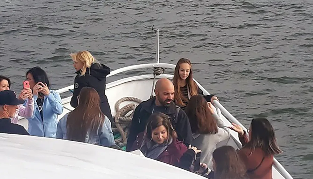 A group of people is gathered on a boat some looking out at the water while others engage with their phones possibly taking photos or videos