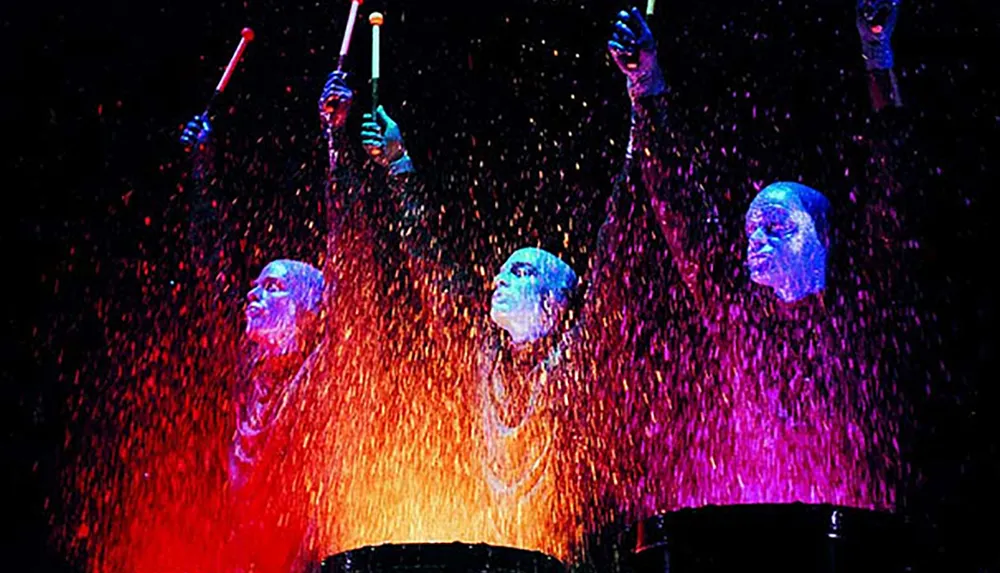 Three performers with blue-painted skin are drumming under a cascade of bright splashes in a dynamic and colorful stage performance