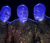 Three performers with blue-painted faces and speckled black shirts appear illuminated under a dark background