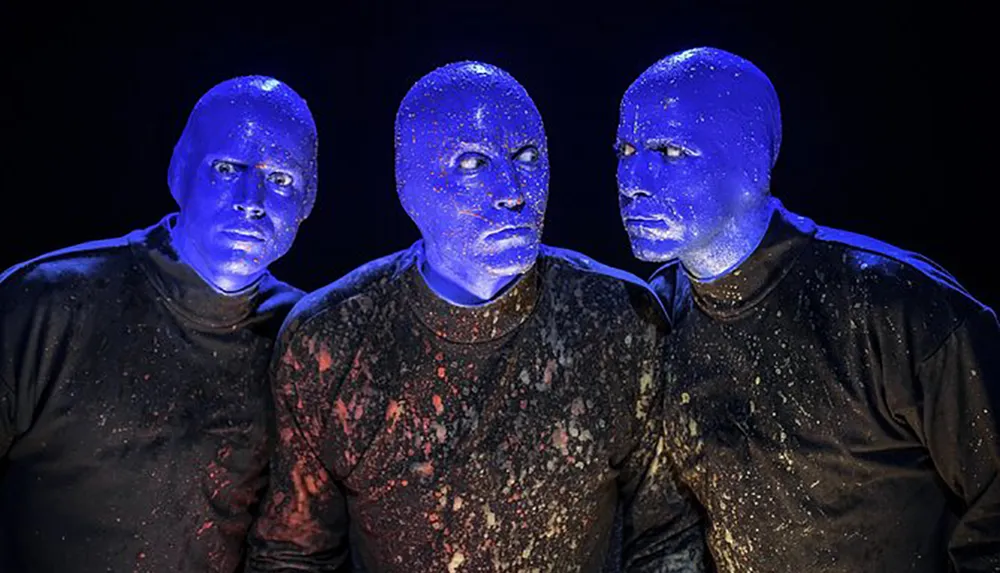 Three performers with blue-painted faces and speckled black shirts appear illuminated under a dark background