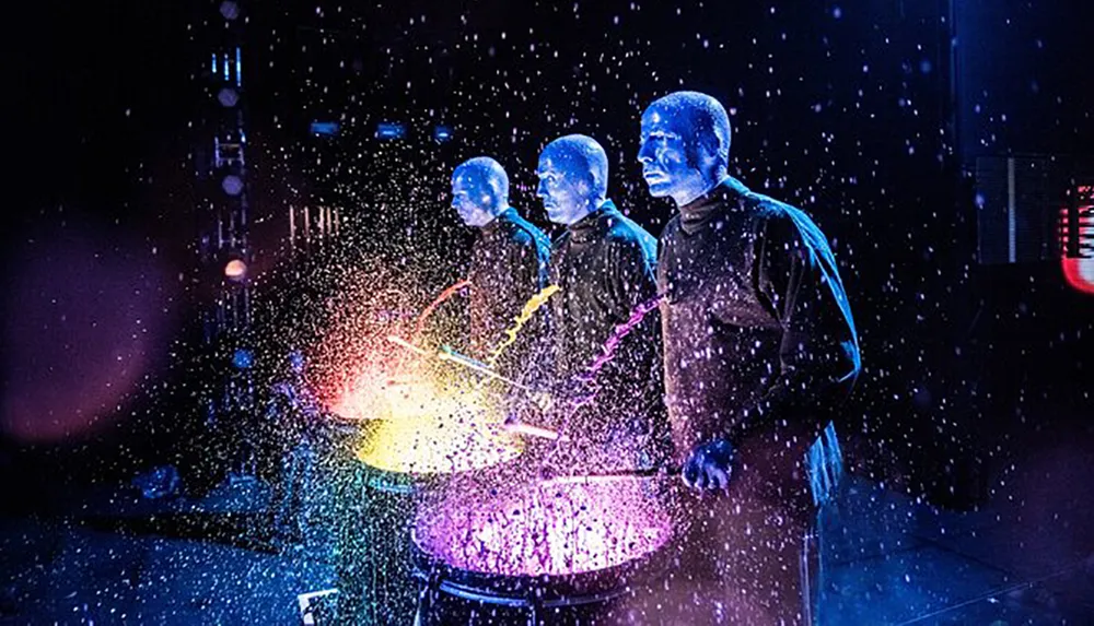 Three performers with blue-painted faces are caught mid-performance amidst a vibrant spray of colors