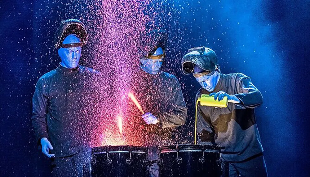 Three performers in blue face paint are interacting with a vibrant spray of particles on a dark stage creating a visually striking scene