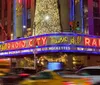 The image displays the vibrant illuminated marquee of Radio City Music Hall at night with blurred traffic in the foreground suggesting the bustling energy of an urban environment