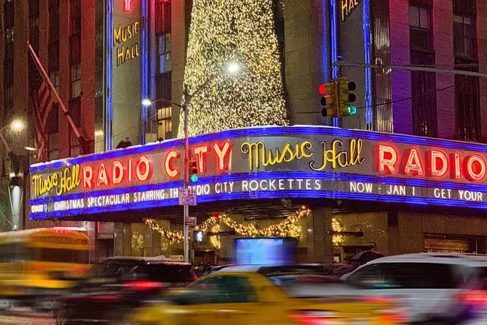 The image displays the vibrant illuminated marquee of Radio City Music Hall at night with blurred traffic in the foreground suggesting the bustling energy of an urban environment