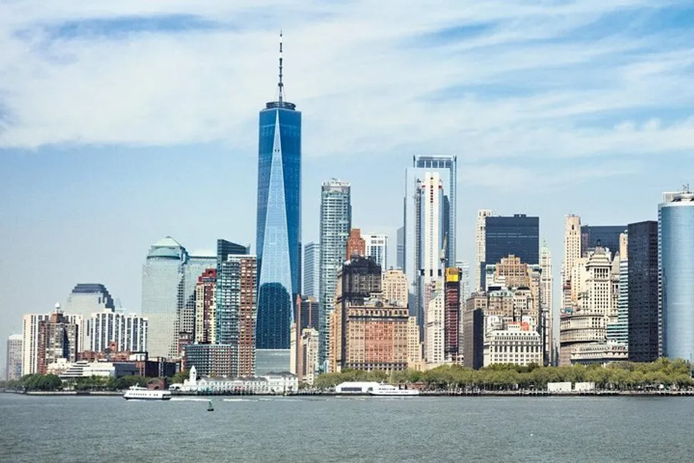 The image shows a view of the Manhattan skyline with the One World Trade Center standing prominently against a blue sky as seen from the water