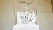 The image shows a large, seated marble sculpture of a historic figure inside a neoclassical monument, accompanied by an inscription above that pays homage to the individual's legacy.