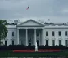 The image shows the White House the official residence of the President of the United States with a green lawn and an American flag flying atop the building on a cloudy day
