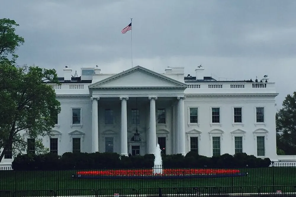 The image shows the White House the official residence of the President of the United States with a green lawn and an American flag flying atop the building on a cloudy day