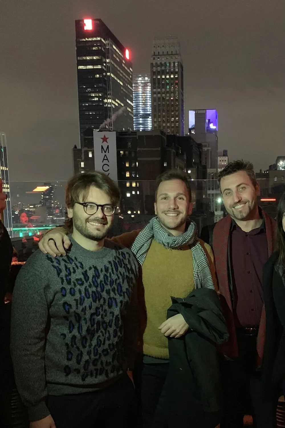 Three men are smiling for a photo on a rooftop at night with illuminated city buildings in the background