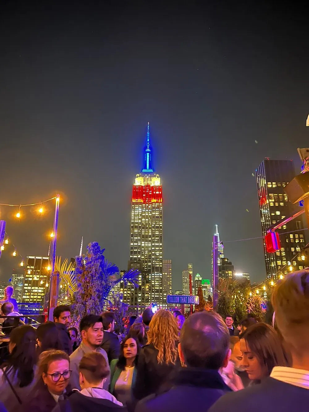 A bustling rooftop gathering with people socializing under the illuminated night sky with the Empire State Building prominent in the background