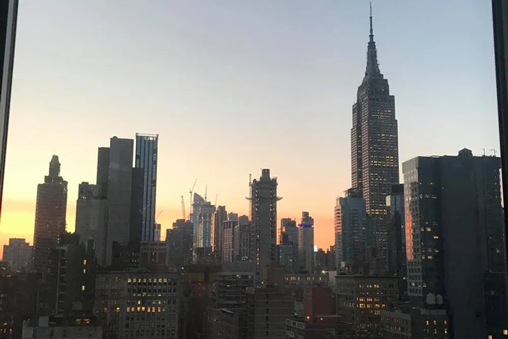 The image shows a skyline view of the Empire State Building amid surrounding buildings during sunset seen through a window