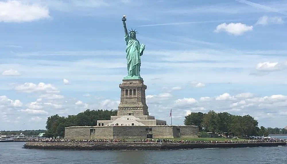 The image shows the Statue of Liberty an iconic symbol of freedom and democracy standing tall on Liberty Island with a clear blue sky and scattered clouds in the background