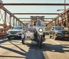 A person riding a motorcycle with a sidecar is moving down a city bridge flanked by cars on one side and with complex metal structures overhead as the passenger turns to smile at the camera