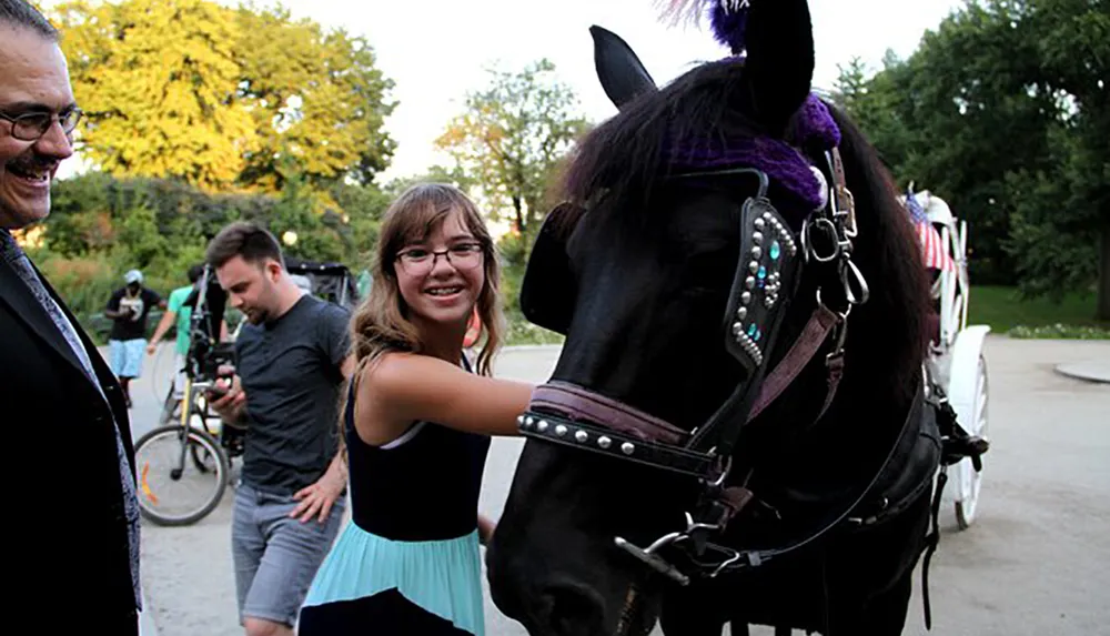 A smiling young woman is petting a horse harnessed to a carriage with a cheerful man looking on and another individual with a bike in the background