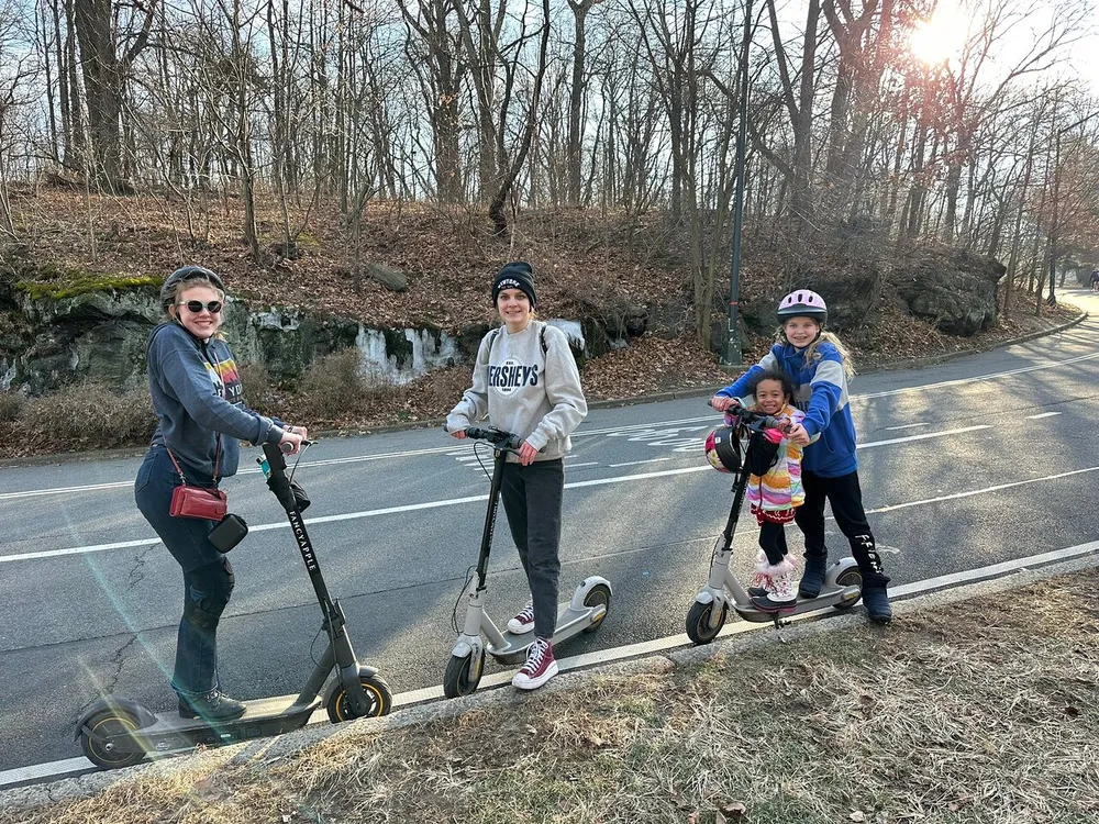Four individuals are happily posing for a photo with their electric scooters on a pathway beside a road with trees and a setting sun in the background