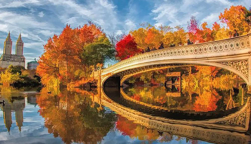 An elegant white bridge arches over a calm body of water reflecting vibrant autumn foliage amidst city buildings in the background