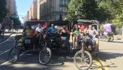 A group of smiling people is enjoying a ride in pedicabs on a sunny city street.