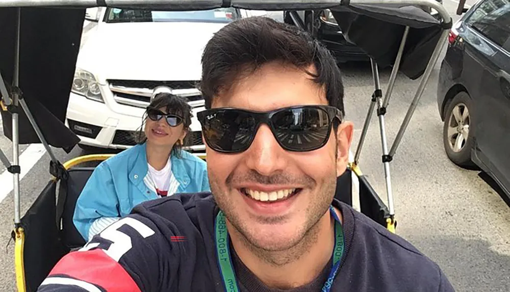 A man in the foreground is smiling for a selfie while a woman in the background sits in a utility vehicle both wearing sunglasses