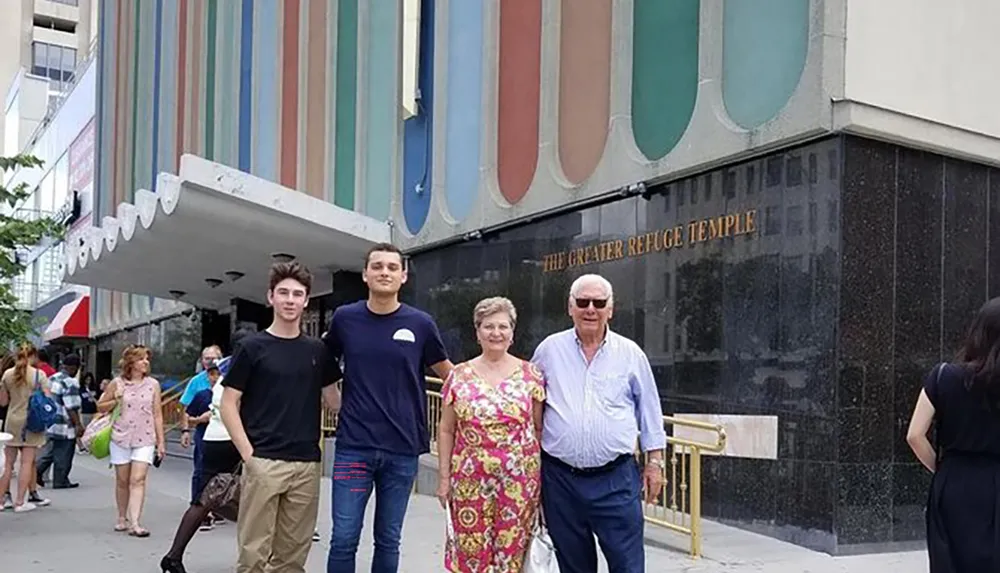 Two younger men and an older couple pose for a photo on a city sidewalk with the entrance of The Greater Refuge Temple building as their backdrop